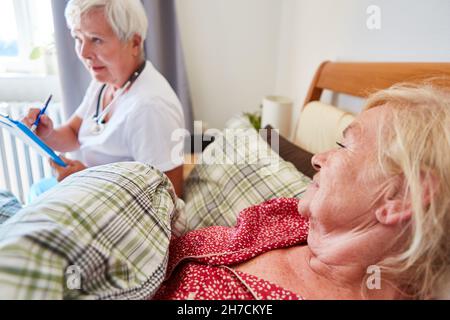 Ältere Menschen als Patient im Bett und ein Arzt mit Patientenakten während eines Hausbesuchs im Pflegeheim Stockfoto