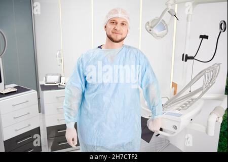 Vorderansicht des bärtigen jungen Stomatologen, der in der modernen Zahnarztpraxis stand und die Kamera ansah und lächelte. Konzept der Zahnmedizin, Stomatologie und Zahnpersonal. Stockfoto