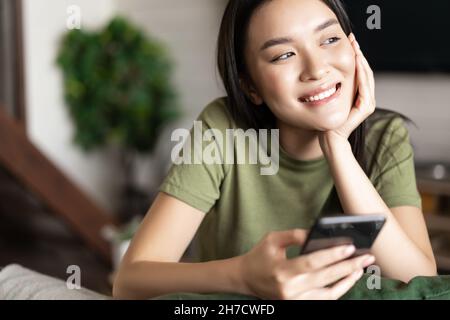 Junge asiatische Frau, die eine Bestellung aufgab, auf die Lieferung über die Handy-App wartete, zu Hause saß und vor dem Fenster schaute Stockfoto