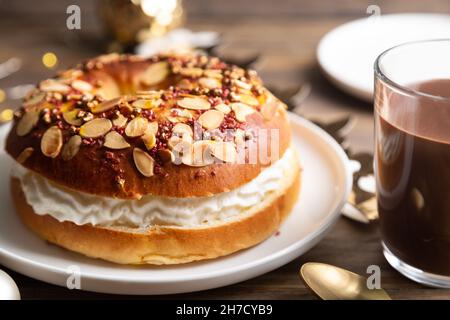 Kleiner 'Roscon de reyes', typisch spanischer Nachtisch von Epiphany, gefüllt mit Schlagsahne und einer heißen Schokoladenschale auf einem rustikalen Holztisch Stockfoto