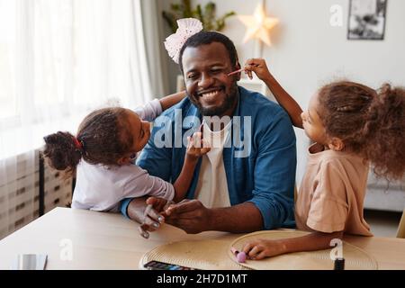 Porträt eines glücklichen afroamerikanischen Vaters, der zu Hause mit Töchtern spielt und sich schminkt Stockfoto