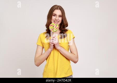 Süße Regenbogenbonbons. Porträt einer positiv braun behaarten Frau im lässigen T-Shirt mit Lollipop und Blick auf die Kamera mit fröhlicher Ausdruckskraft. Innenaufnahme des Studios isoliert auf grauem Hintergrund. Stockfoto