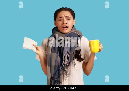 Ungesunde kranke Frau mit schwarzen Dreadlocks, trägt weißes Hemd und ist in einen Schal gewickelt, hält eine Tasse Tee, hat Grippe und laufende Nase. Innenaufnahme des Studios isoliert auf blauem Hintergrund. Stockfoto