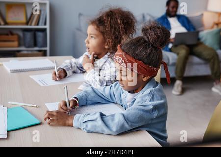 Seitenansicht des Porträts von zwei afroamerikanischen Mädchen, die Hausaufgaben machen, während sie am Schreibtisch in einem gemütlichen Zuhause sitzen Stockfoto