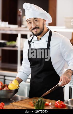Positiver asiatischer Koch hält Messer in der Nähe von frischem Gemüse und Schneidebrett in der Küche Stockfoto