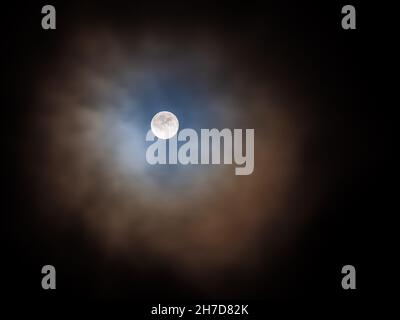 Szenische Ansicht des Mondes am 19.11.2021 vor der Mondfinsternis. Der Mond scheint durch einen wolkigen Himmel mit einem blauen und orangen Schein, der ihn umgibt. Stockfoto