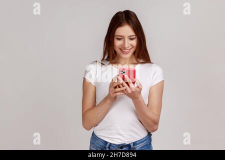 Glückliche Frau mit Mobiltelefon und lächelt freudig, tippen oder lesen lustige Nachricht in sozialen Netzwerken, Online-Kommunikation, trägt weißes T-Shirt. Innenaufnahme des Studios isoliert auf grauem Hintergrund. Stockfoto