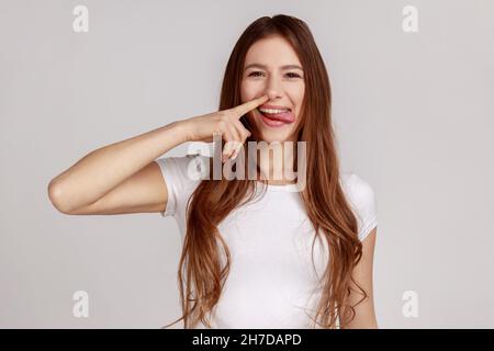 Lustige fröhliche Frau, die Nase pflückt mit dummen dummen, dummen Gesichtern, ausziehenden Boogern, schlechten Manieren Konzept, Fehlverhalten, trägt weißes T-Shirt. Innenaufnahme des Studios isoliert auf grauem Hintergrund. Stockfoto