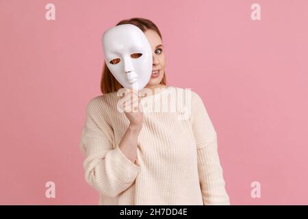 Portrait einer lächelnden blonden Frau, die weiße Maske hält, guckt, die Kamera anschaut, vorgibt, eine andere Person zu sein, trägt weißen Pullover. Innenaufnahme des Studios isoliert auf rosa Hintergrund. Stockfoto