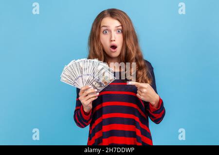 Schockierte Frau, die einen gestreiften Pullover im lässigen Stil trägt, auf Dollarnoten in der Hand zeigt, mit erstauntem Ausdruck und großem Gewinn. Innenaufnahme des Studios isoliert auf blauem Hintergrund. Stockfoto