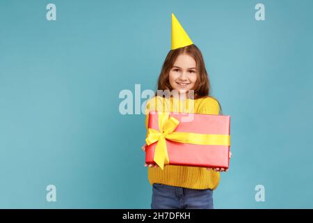 Portrait des kleinen Mädchens in Party-Kegel geben Geschenk-Box, gratuliert ihrem Freund mit Geburtstag, tragen gelben lässigen Stil Pullover. Innenaufnahme des Studios isoliert auf blauem Hintergrund. Stockfoto