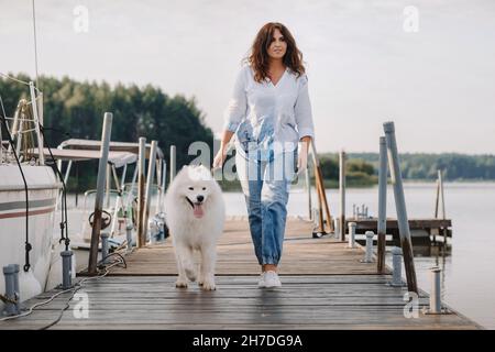 Eine glückliche Frau mit einem großen weißen Hund spaziert auf dem Pier in der Nähe der Yacht und des Meeres. Stockfoto