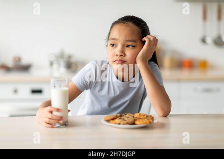 Porträt von traurigen asiatischen Mädchen hält Glas Milch und träumen, wegschauen, während in der Küche Innenraum sitzen Stockfoto