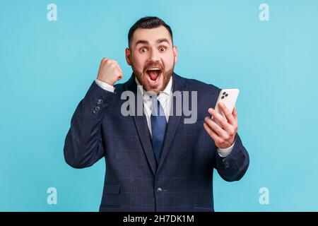 Ich bin Champion. Attraktiver Mann mit Bart trägt dunklen offiziellen Stil Anzug stehen, halten Telefon, machen Selfie und feiern den Sieg. Innenaufnahme des Studios isoliert auf blauem Hintergrund. Stockfoto