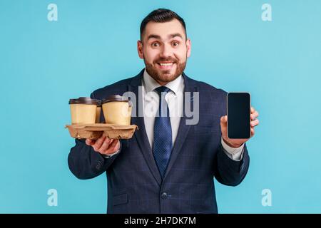 Extrem glücklich Mann trägt offiziellen Stil Anzug in den Händen halten leeren Display Smartphone und Halter mit Kaffee Pappbecher, Online-Bestellung. Innenaufnahme des Studios isoliert auf blauem Hintergrund. Stockfoto