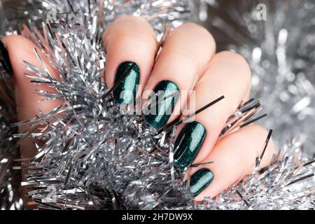 Weibliche Hand mit schönen Urlaub Maniküre - grün glitzernden Nägeln mit silbernen Weihnachten Lametta Hintergrund. Selektiver Fokus. Nahaufnahme. Nagelpflege Stockfoto