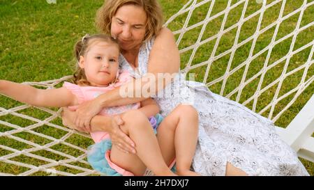 Das fröhliche Paar Großmutter und Enkelin 3 Jahre alt entspannen sich und sonnen sich in einer Hängematte auf einer grünen Wiese in ihrem Garten an einem sonnigen Sommer Stockfoto
