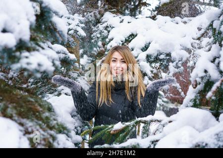 Portrait schönes Mädchen im Winter verschneiten Tag in der Nähe von Tannenbaum im Wald. Junge lächelnde Frau, die mit Schnee in Fäustlingen spielt. Das Weibchen genießt die Natur und atmet saubere, frostige Luft in warmer Kleidung. Winterkonzept Stockfoto