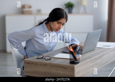 Müde verärgert traurige tausendjährige indische Frau, die an Rückenschmerzen am Arbeitsplatz im Büroinneren leidet Stockfoto