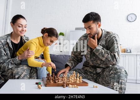 Nachdenklicher Vater in Tarnuniform spielt Schach in der Nähe lächelnder Mutter und Kind zu Hause Stockfoto