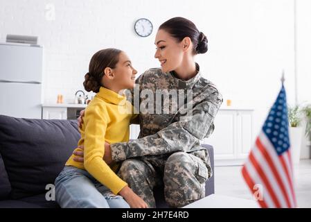 Seitenansicht der Mutter in Militäruniform, die das Kind in der Nähe der amerikanischen Flagge auf dem Tisch zu Hause umarmt Stockfoto
