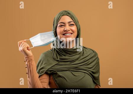 Ende der Epidemie. Glückliche arabische Frau, die ihre Gesichtsmaske auf braunem Studiohintergrund auszieht. Coronavirus-Wiederherstellungskonzept Stockfoto