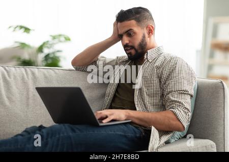 Schockierter, tausendjähriger arabischer Mann, der seinen Kopf vor den Laptop schnappte und Fehler beim Online-Arbeitsprojekt im Heimbüro machte Stockfoto