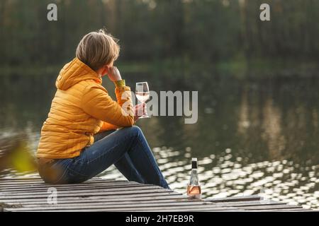 Frau in einer gelben Jacke, die sich auf einem hölzernen Pier auf einem See mit einem Glas Rosenwein entspannt und den Sonnenuntergang im Herbst alleine beobachtet. Genießen Sie die Natur, Entspannung und med Stockfoto