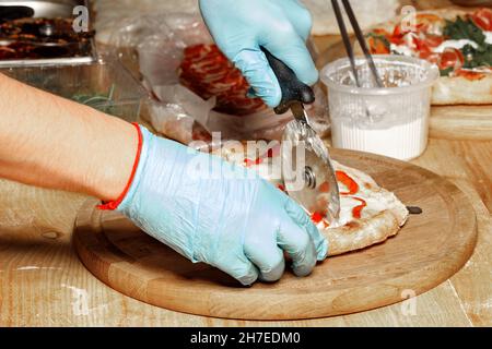 Die Hände eines Küchenchefs, eines kaukasischen Mannes in blauen Latexhandschuhen, schneiden die gekochte Pizza auf einem hölzernen Küchenbrett. Nahaufnahme. Stockfoto