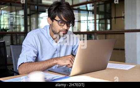 Ein junger indischer Geschäftsmann schaut sich das Webinar im Büro auf einem Laptop an. Stockfoto