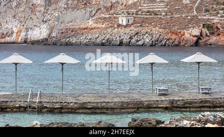 Gerolimenas Dorf bei Mani Laconia Peloponnes Griechenland. Steile Klippen Stein gepflasterten Plattform mit Sonnenschirmen und Liegen zum Entspannen in ruhigen blauen se Stockfoto