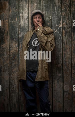 Junkie-Mann mit Mütze grüner Anorak und blauer Jogginghose in der Nähe einer alten hölzernen Wand Stockfoto