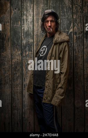 Junkie-Mann mit Mütze grüner Anorak und blauer Jogginghose in der Nähe einer alten hölzernen Wand Stockfoto