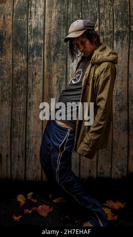 Junkie-Mann mit Mütze grüner Anorak und blauer Jogginghose in der Nähe einer alten hölzernen Wand Stockfoto