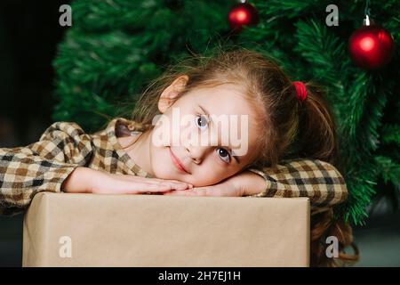 Nettes Mädchen, das ihren Kopf auf ihre Hände auf eine eingewickelte Geschenk-Box legte und posierte. Sitzt unter einem weihnachtsbaum, der mit roten glänzenden Kugeln geschmückt ist. Stockfoto