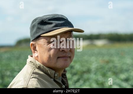 Porträt eines ernsthaft gealterten asiatischen Bauern in Kappe, der am Sommertag auf dem Kohlplantage-Feld steht Stockfoto
