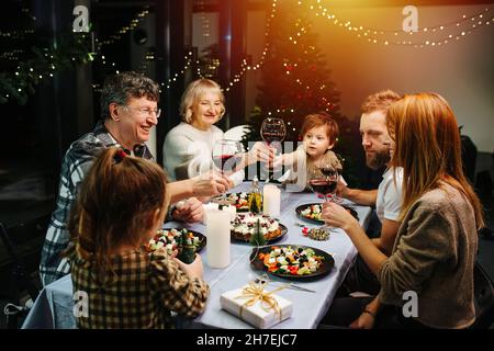 Lebhafte große Familie zum weihnachtsfest. Sitzen um einen großen Esstisch, klirren Weingläser, feiern. Weihnachtsbaum und Girlanden im Hintergrund Stockfoto