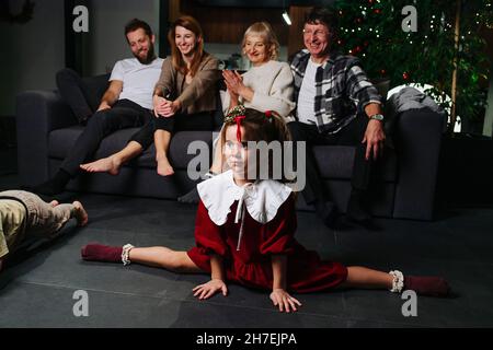 Etwas gelangweilt kleines Mädchen in rotem Kleid sitzt in den Splits. Ihre große Familie sitzt auf einem Sofa im Hintergrund zusammen mit weihnachtsbaum. Stockfoto