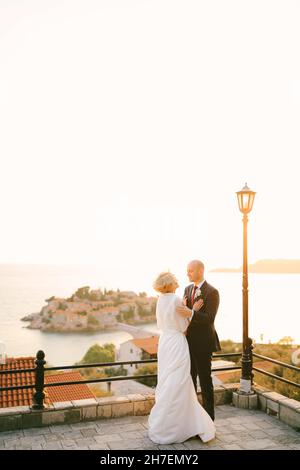 Der Bräutigam umarmt die Schultern der Braut in einem weißen Kleid und Umhang, während er auf der Aussichtsplattform mit Blick auf die Insel Sveti Stefan in der Nähe von Budva steht Stockfoto