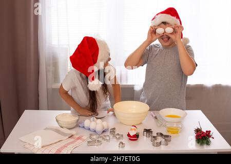 Bruder und Schwester in roten Mützen haben Spaß und bereiten Weihnachtskekse an einem weißen Tisch zu. Stockfoto