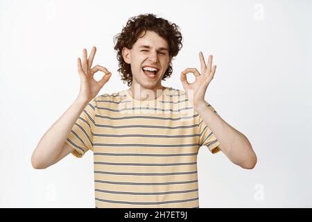 Glücklicher, gutaussehender junger Mann, der ein gutes Zeichen zeigt, lachend und vor Zustimmung nickt und über weißem Hintergrund steht Stockfoto