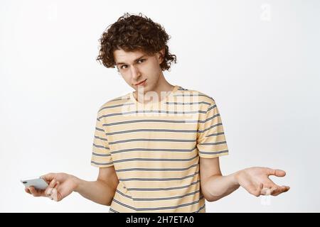 Verwirrt junger nerdy Kerl, zuckende Schultern und halten Handy, Int T-Shirt auf weißem Hintergrund stehen Stockfoto