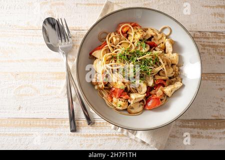 Vollkorn-Spaghetti mit Tomaten-, Pilz- und Hähnchenstreifen, garniert mit Kresse, grauem Teller und Besteck auf einem hellen rustikalen Holztisch, Draufsicht Stockfoto