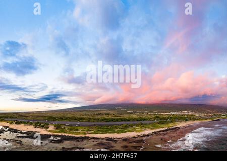 Ein schwacher Regenbogen bildet sich, während die untergehende Sonne rosa Wolken über den Hängen des Hualalai Vulkans aufleuchtet, wie sie vom Old Kona Airport County Park, Hawaii, aus gesehen werden Stockfoto