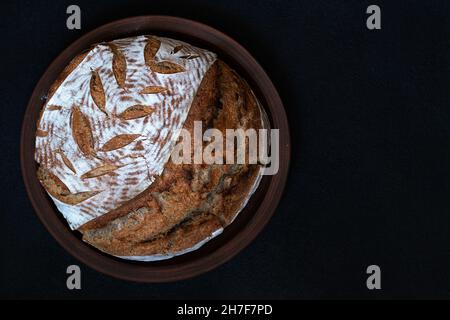 Hausgemachtes rundes dunkles Brot mit Mehlkruste und bemaltem Stachelett auf einem Tonteller. Draufsicht mit Kopierbereich. Stockfoto