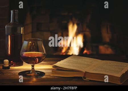 Ein Glas Cognac, eine Flasche und ein offenes altes Buch auf dem Tisch in der Nähe des brennenden Kamins. Ruhe- und Entspannungskonzept Stockfoto