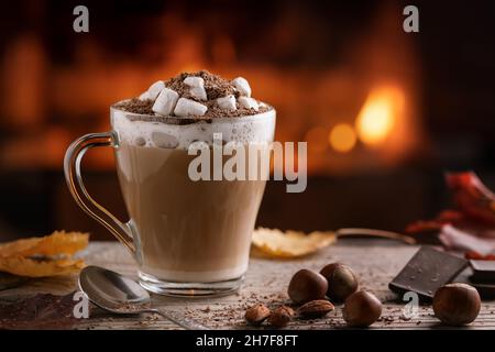 Kakao mit Salmellchen und Schokolade in einem Glaskrug auf einem Holztisch in der Nähe eines brennenden Kamins. Stockfoto