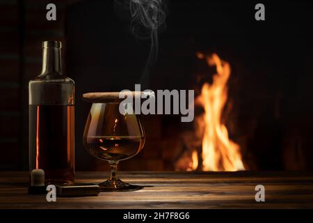 Ein Glas Cognac, eine Zigarre, eine Flasche auf dem Tisch in der Nähe des brennenden Kamins. Entspannungs- und Genusskonzept Stockfoto