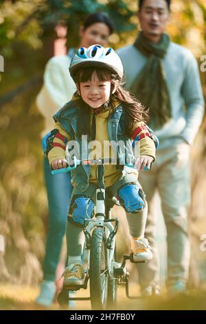 Kleine asiatische Mädchen mit Helm und voller Schutz Zahnräder Reiten Fahrrad im Stadtpark mit Eltern beobachten von hinten Stockfoto