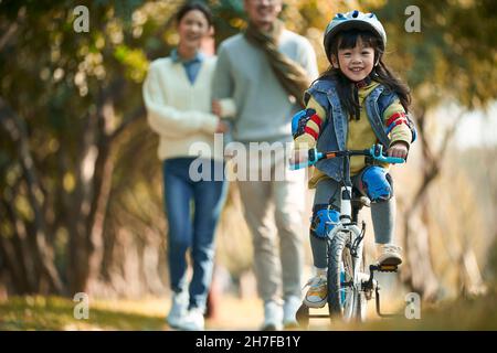 Kleine asiatische Mädchen mit Helm und voller Schutz Zahnräder Reiten Fahrrad im Stadtpark mit Eltern beobachten von hinten Stockfoto